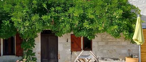 Un bâtiment rustique avec une porte en bois, une fenêtre avec des volets et un petit espace extérieur aménagé avec une table et des chaises.