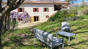 Una casa in pietra con tetto in tegole, un patio con tavolo e sedie, e un pendio erboso.