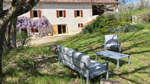 Una casa de pedra amb teulada d'àmples rajoles, un pati amb taula i cadires, i una pendent coberta d'herba.