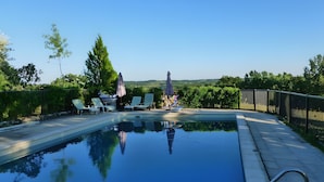 Una piscina rectangular amb una escala metàl·lica, envoltada de gandules i ombrelles, i vista a zones verdes i turons al fons.