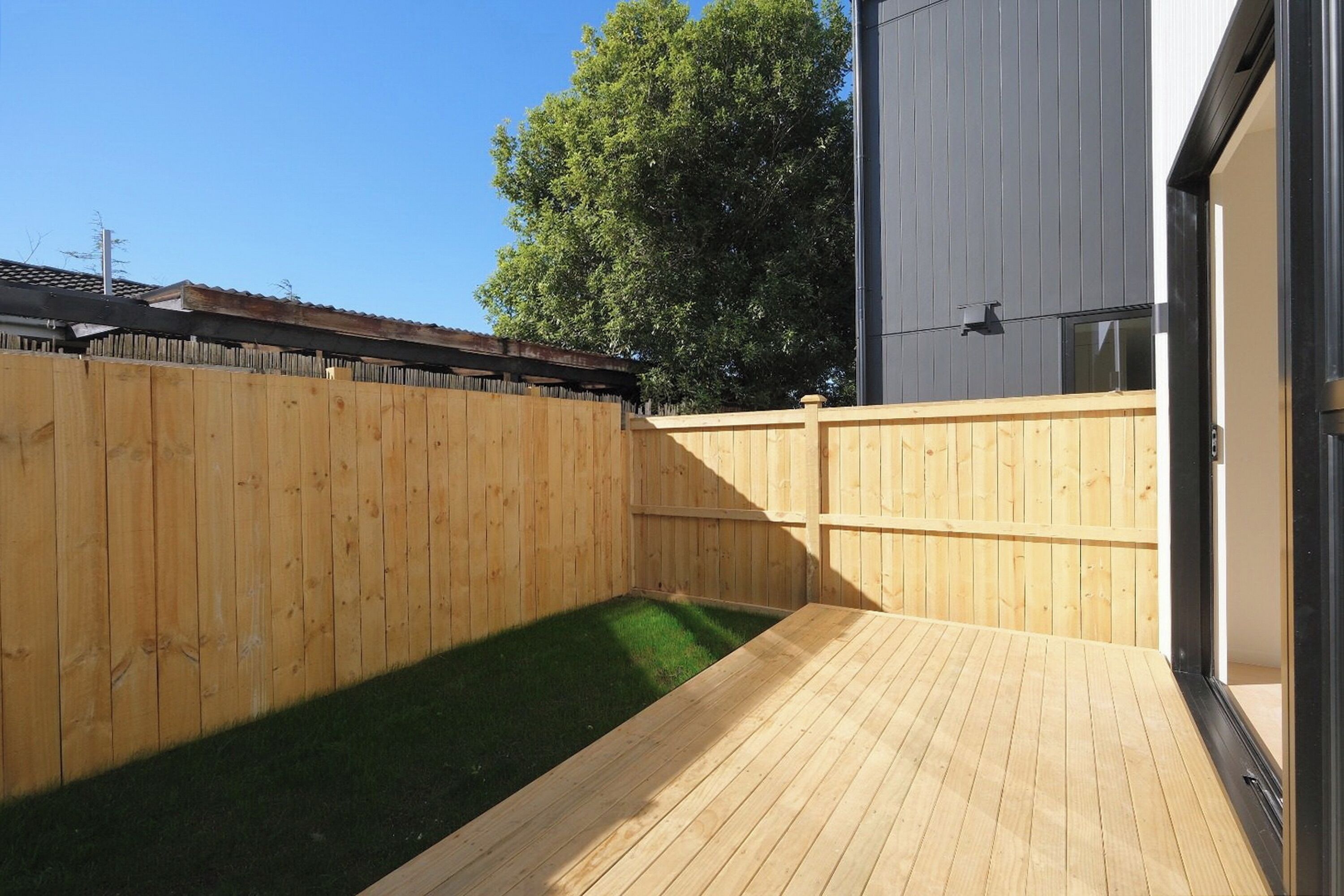 A wooden deck with a small grassy area, a wooden fence, and a building with a metal exterior.