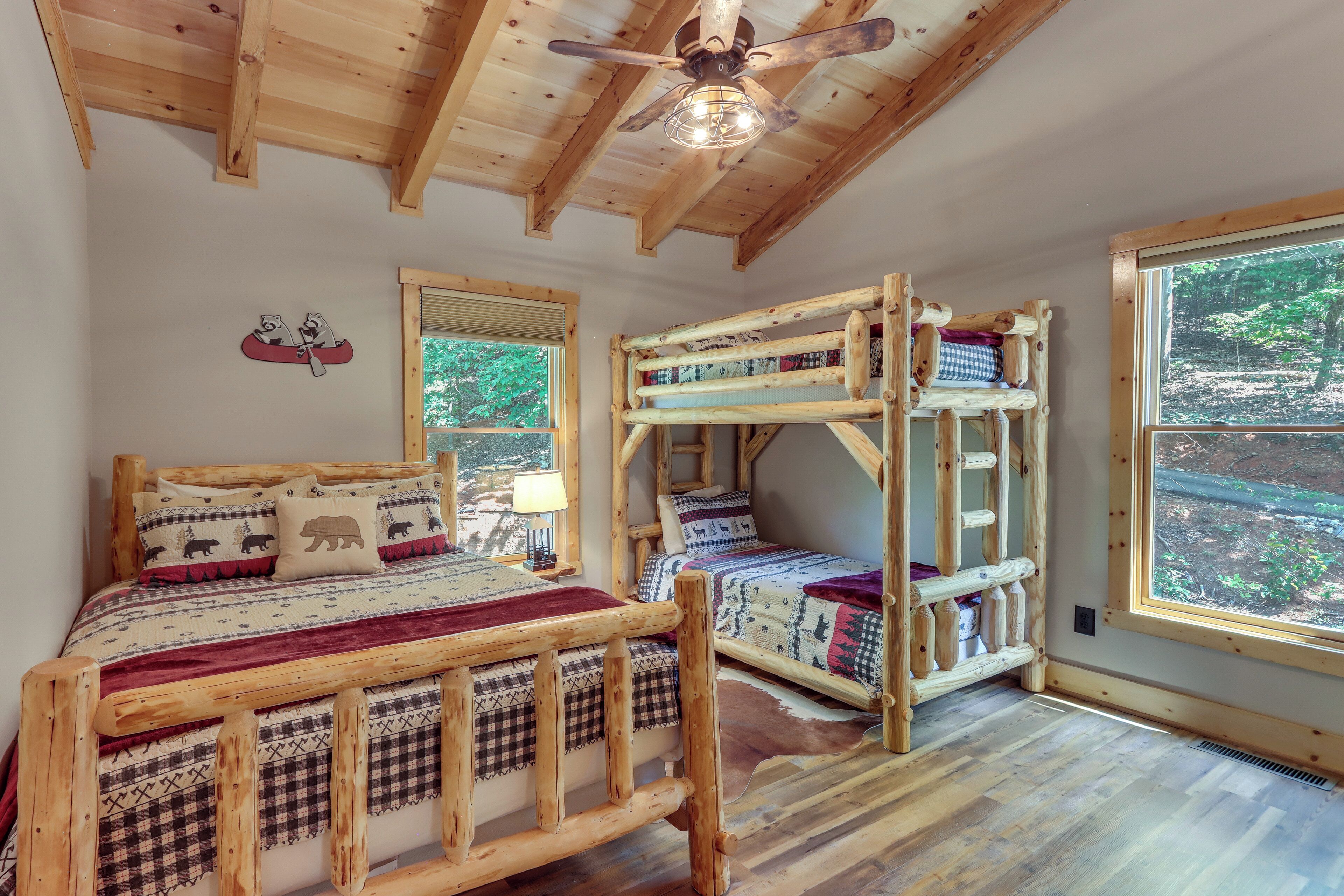 Een slaapkamer in chaletstijl met een houten plafond, een bed met een rode deken, een stapelbed en een raam met uitzicht op een bos.
