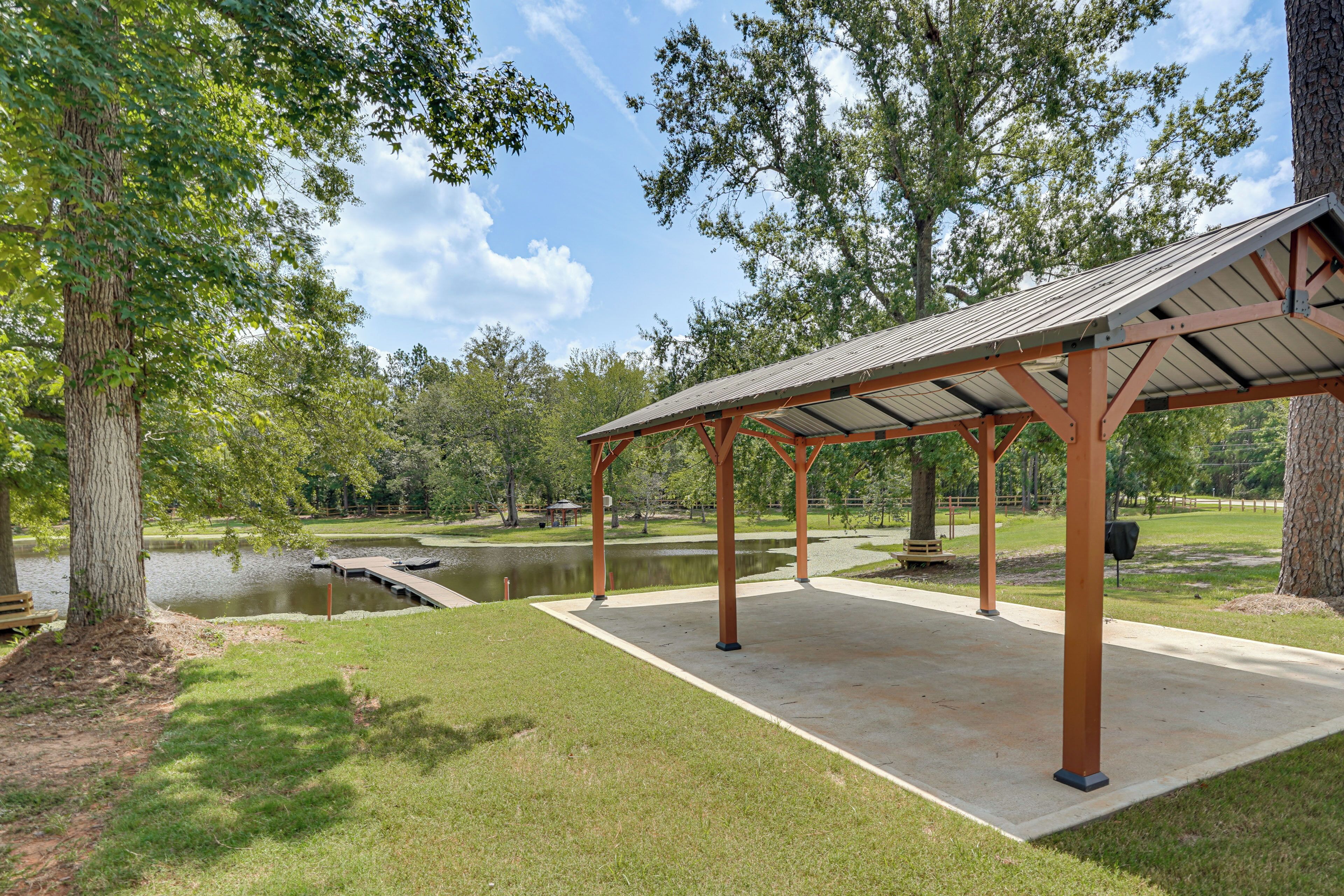 A covered pavilion with a concrete floor, surrounded by grass and trees, near a body of water.