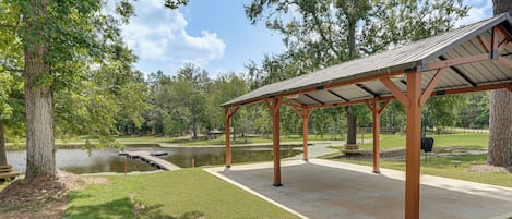 A covered pavilion with a concrete floor, surrounded by grass and trees, near a body of water.