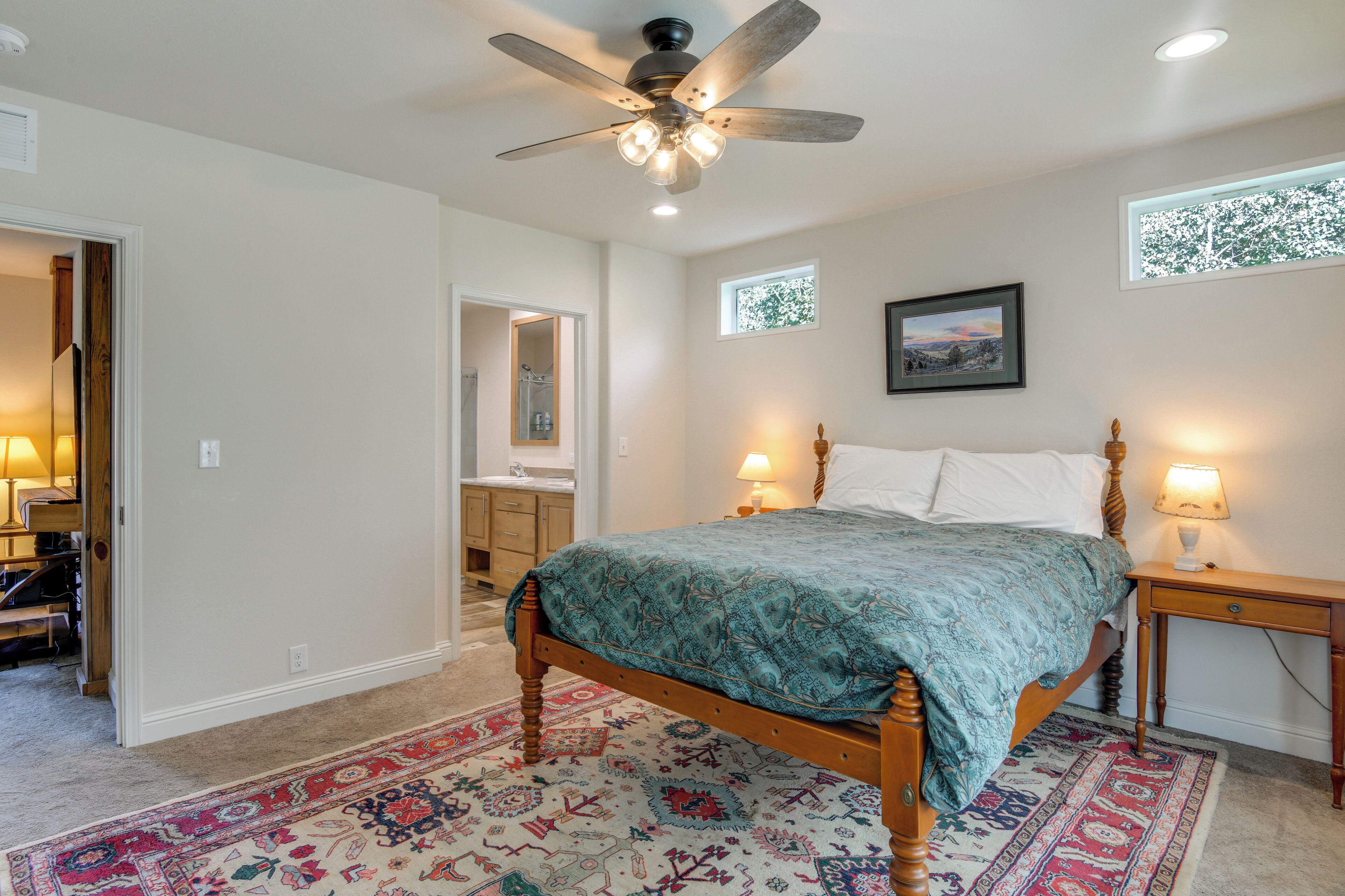 Une chambre à coucher avec un lit en bois, un ventilateur de plafond, une table de chevet, une lampe de chevet, une fenêtre avec vue et une salle de bain visible à travers une porte ouverte.