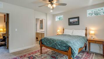 Une chambre à coucher avec un lit en bois, un ventilateur de plafond, une table de chevet, une lampe de chevet, une fenêtre avec vue et une salle de bain visible à travers une porte ouverte.