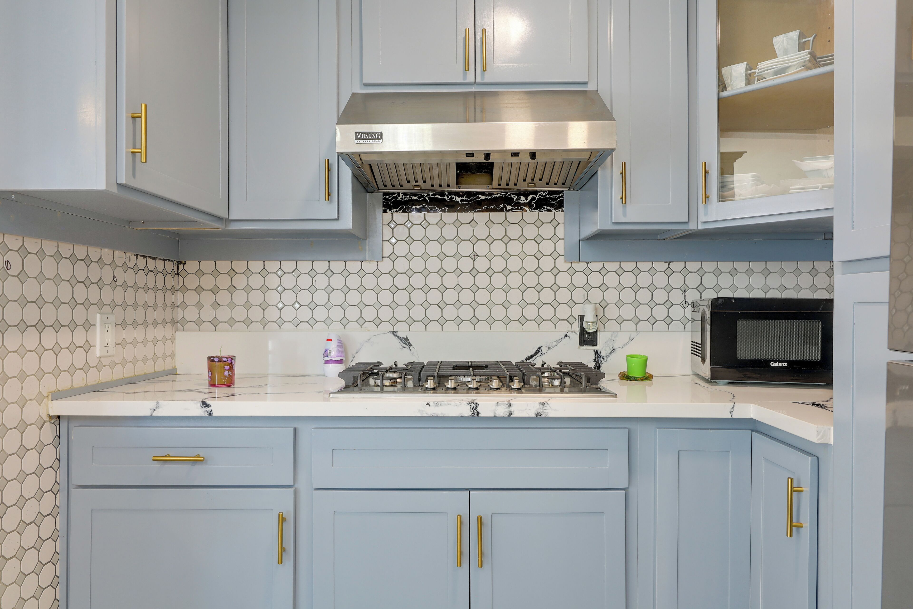 A kitchen with light blue cabinets, a stainless steel range hood, a microwave, and a hexagonal tile backsplash.
