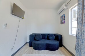A room with a dark blue sofa, a flat-screen TV mounted on the wall, an air conditioning unit, and a window with curtains. - Walkable Location! Spacious Huntington Park Home (Huntington Park)