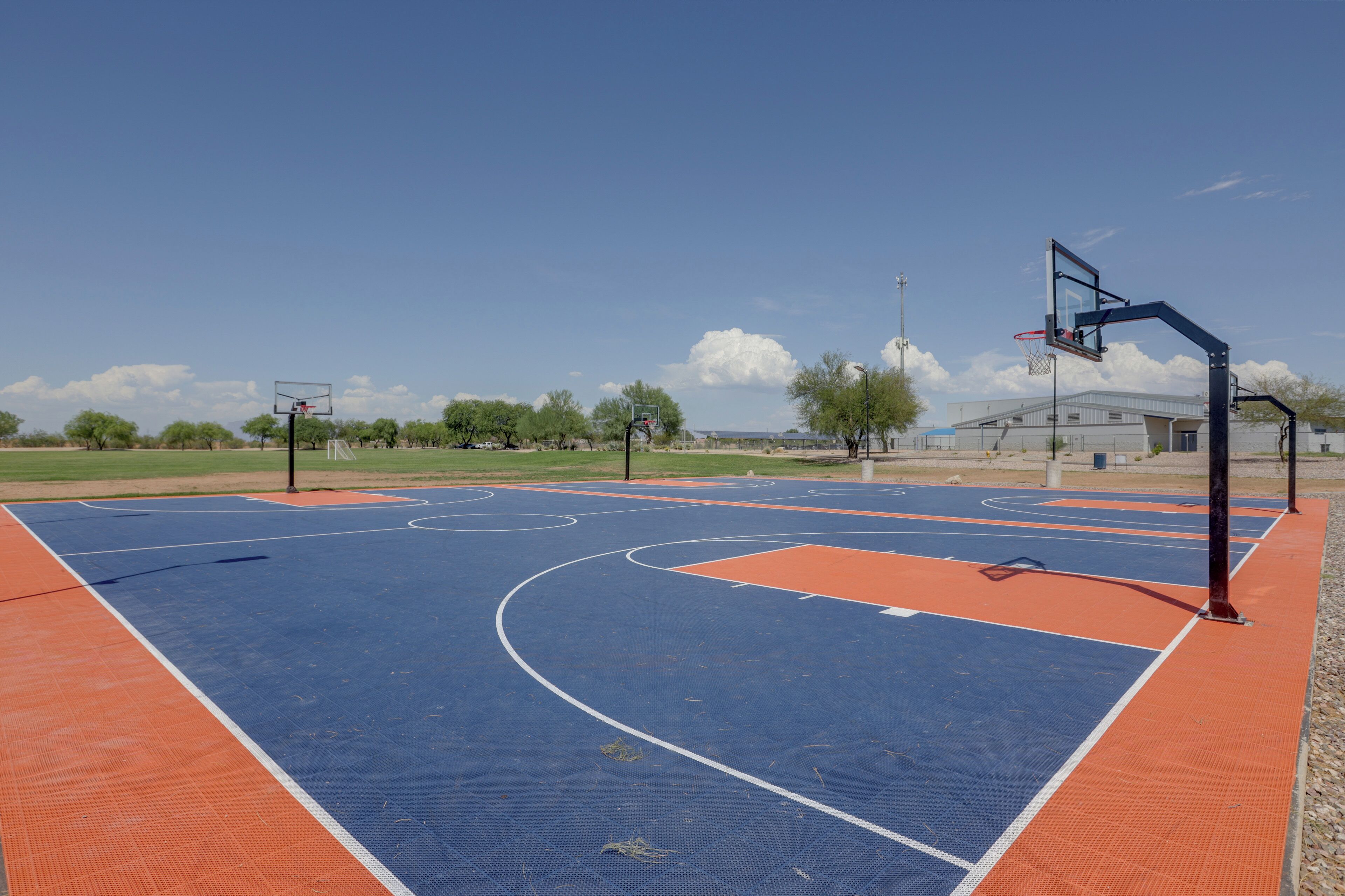 An outdoor basketball court with a blue and orange playing surface, surrounded by trees and a building in the background.