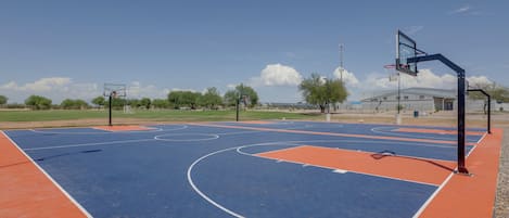 An outdoor basketball court with a blue and orange playing surface, surrounded by trees and a building in the background.