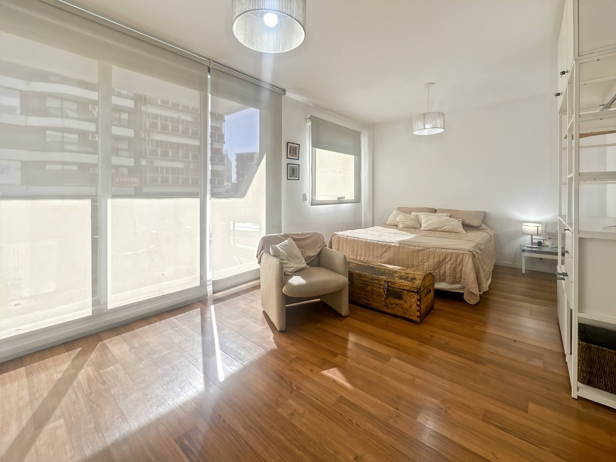 A bedroom with a bed, a chair, a chest of drawers, a bedside table, and a large window.