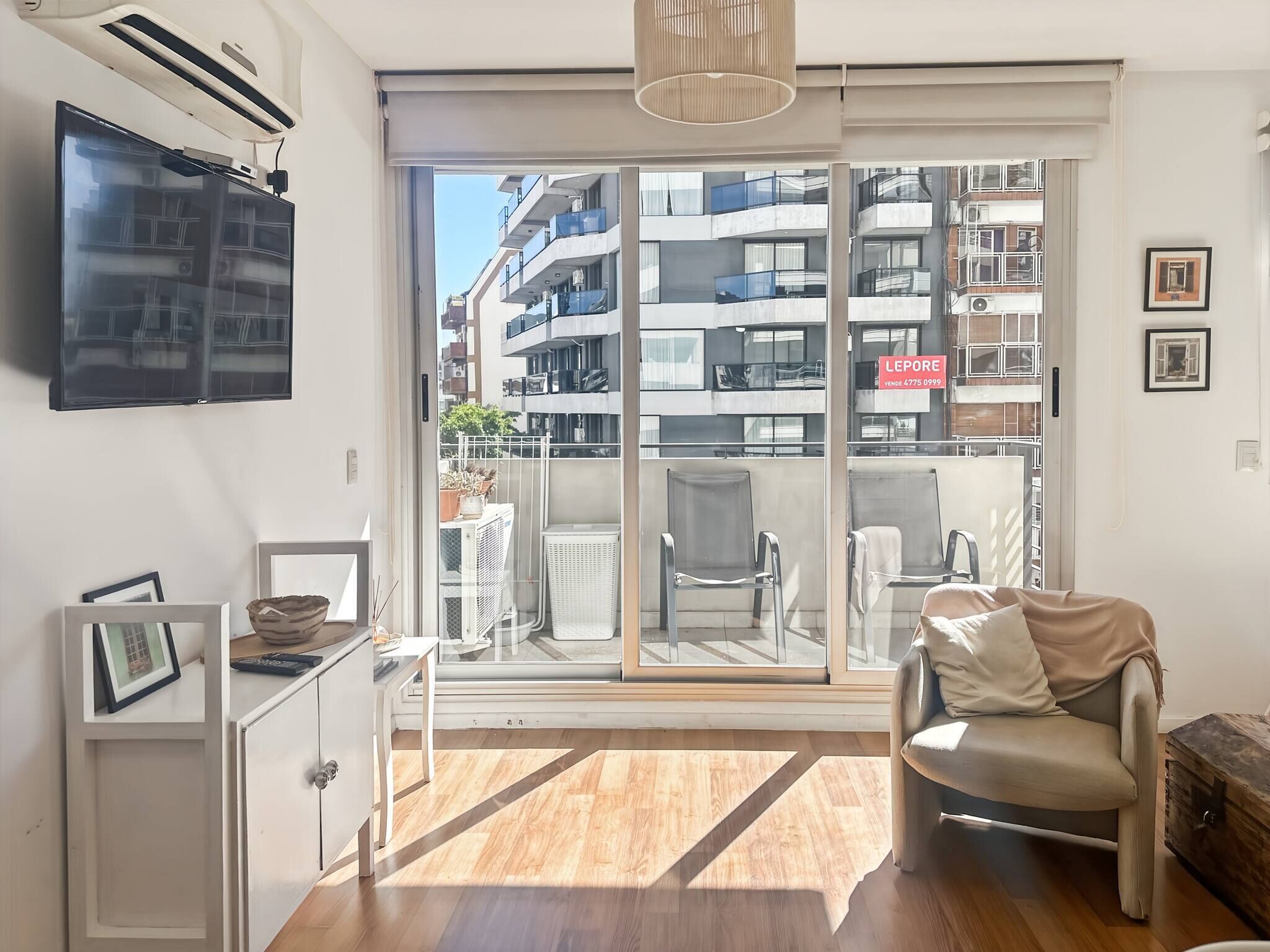 A modern apartment with a large sliding glass door leading to a balcony, a flat-screen TV mounted on the wall, and a minimalist furniture arrangement.