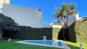 Una piscina rodeada de césped y setos bien cuidados, con una palmera y un edificio al fondo.