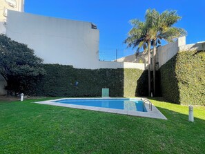 Una piscina rodeada de césped y setos bien cuidados, con una palmera y un edificio al fondo.