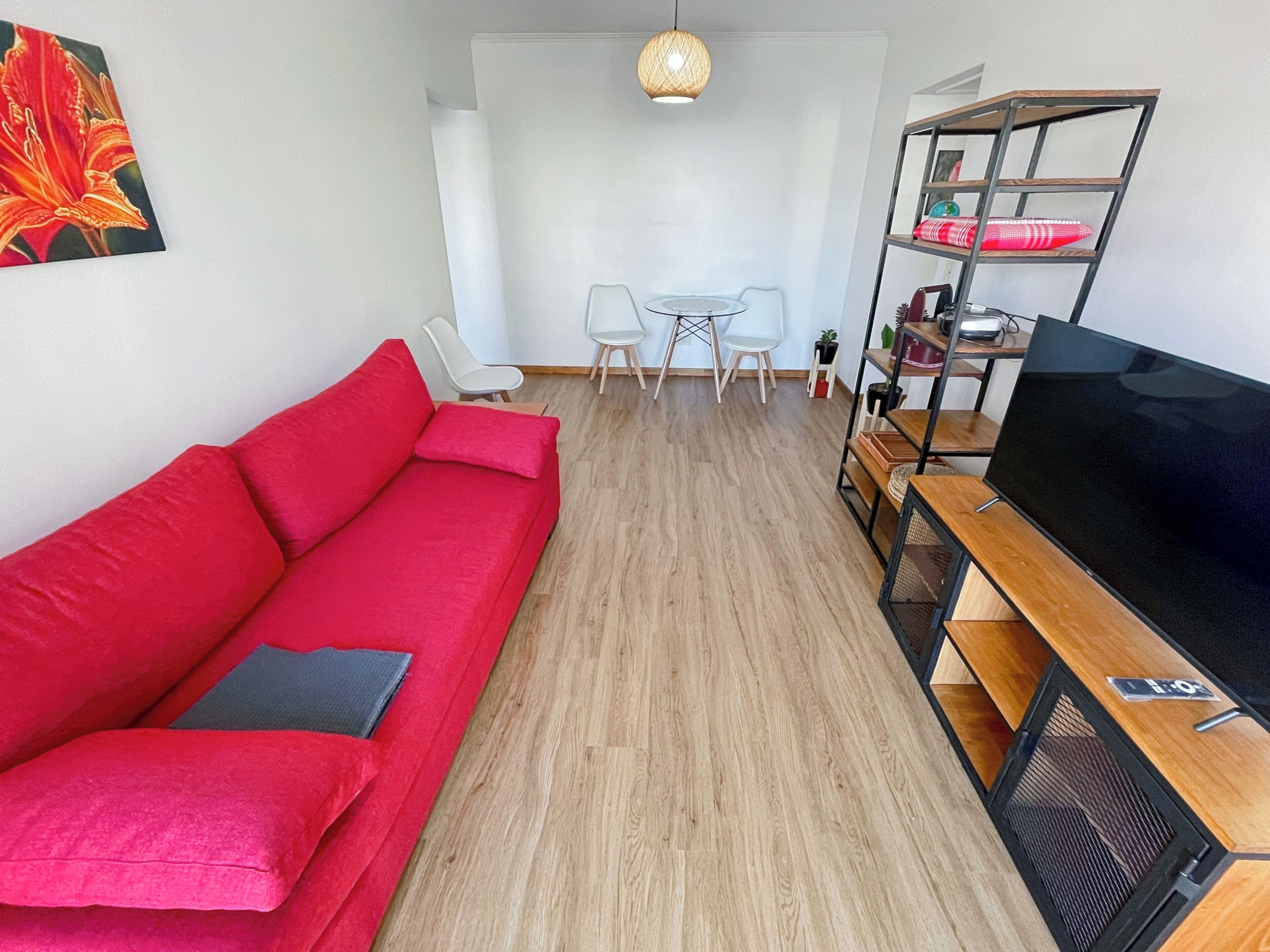 Una sala moderna con un sofá rojo, un televisor sobre un mueble de madera, una zona de comedor con una mesa y sillas, y una lámpara colgante.