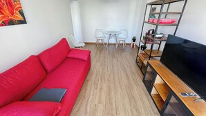 Una sala moderna con un sofá rojo, un televisor sobre un mueble de madera, una zona de comedor con una mesa y sillas, y una lámpara colgante.
