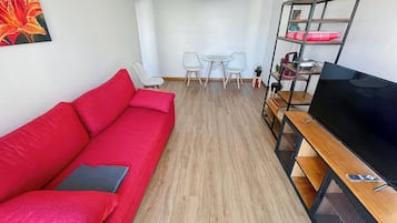 Una sala moderna con un sofá rojo, un televisor sobre un mueble de madera, una zona de comedor con una mesa y sillas, y una lámpara colgante.
