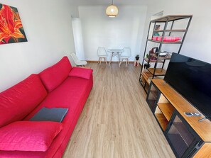 Una sala moderna con un sofá rojo, un televisor sobre un mueble de madera, una zona de comedor con una mesa y sillas, y una lámpara colgante.