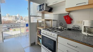 Cocina moderna con estufa a gas, horno, microondas y cafetera. Se ve un letrero que indica lavandería, una vista de la ciudad desde el balcón y un guante de horno rojo colgado en la pared.