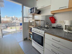 Cocina moderna con estufa a gas, horno, microondas y cafetera. Se ve un letrero que indica lavandería, una vista de la ciudad desde el balcón y un guante de horno rojo colgado en la pared.
