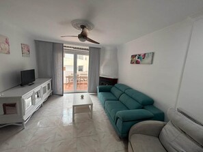 A living room with a white cabinet, a teal sofa, a grey sofa, a ceiling fan, and a sliding glass door leading to an outdoor area. - ResbalajeHomes Granada 75 1A (Nerja)