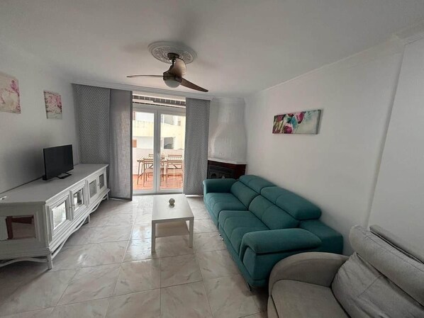 Un salon comprenant un meuble blanc, un canapé turquoise, un canapé gris, un ventilateur de plafond et une porte-fenêtre coulissante donnant sur un espace extérieur.
