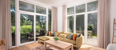 Un salon spacieux avec un grand canapé, une table basse et une vue sur un jardin grâce à de grandes fenêtres du sol au plafond.