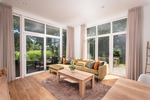 A spacious living room with a large sofa, a coffee table, and a view of a garden through floor-to-ceiling windows. - Stunning Riverside Cabin with Hot Tub (Zuna)