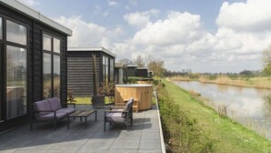 A modern outdoor patio with seating and a hot tub, overlooking a calm water body and a grassy field.