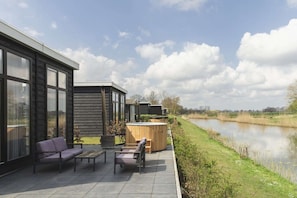 A modern outdoor patio with seating and a hot tub, overlooking a calm water body and a grassy field.