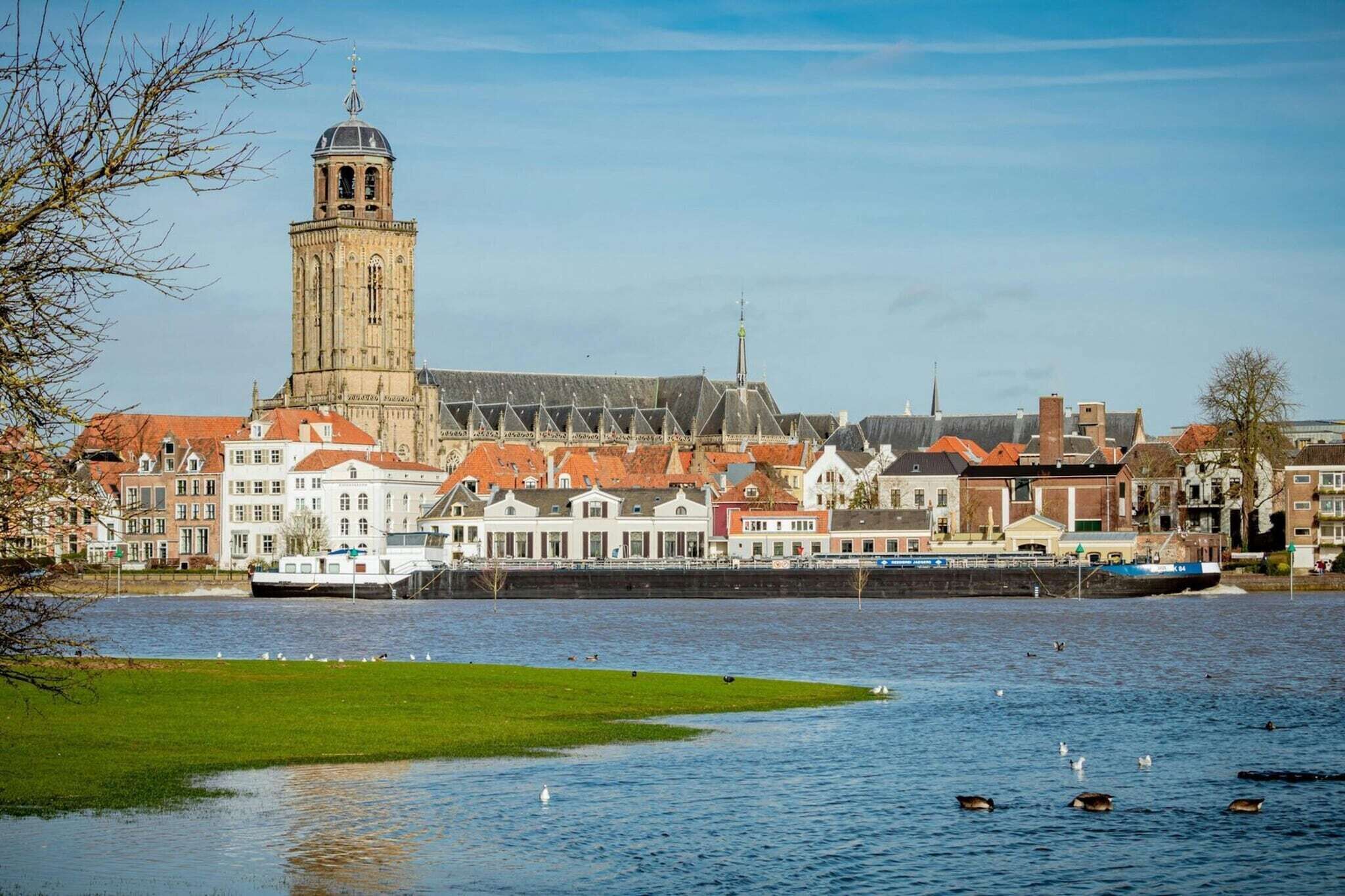 A riverside view of a town with historic buildings, a prominent tower, and a boat docked near the shore.