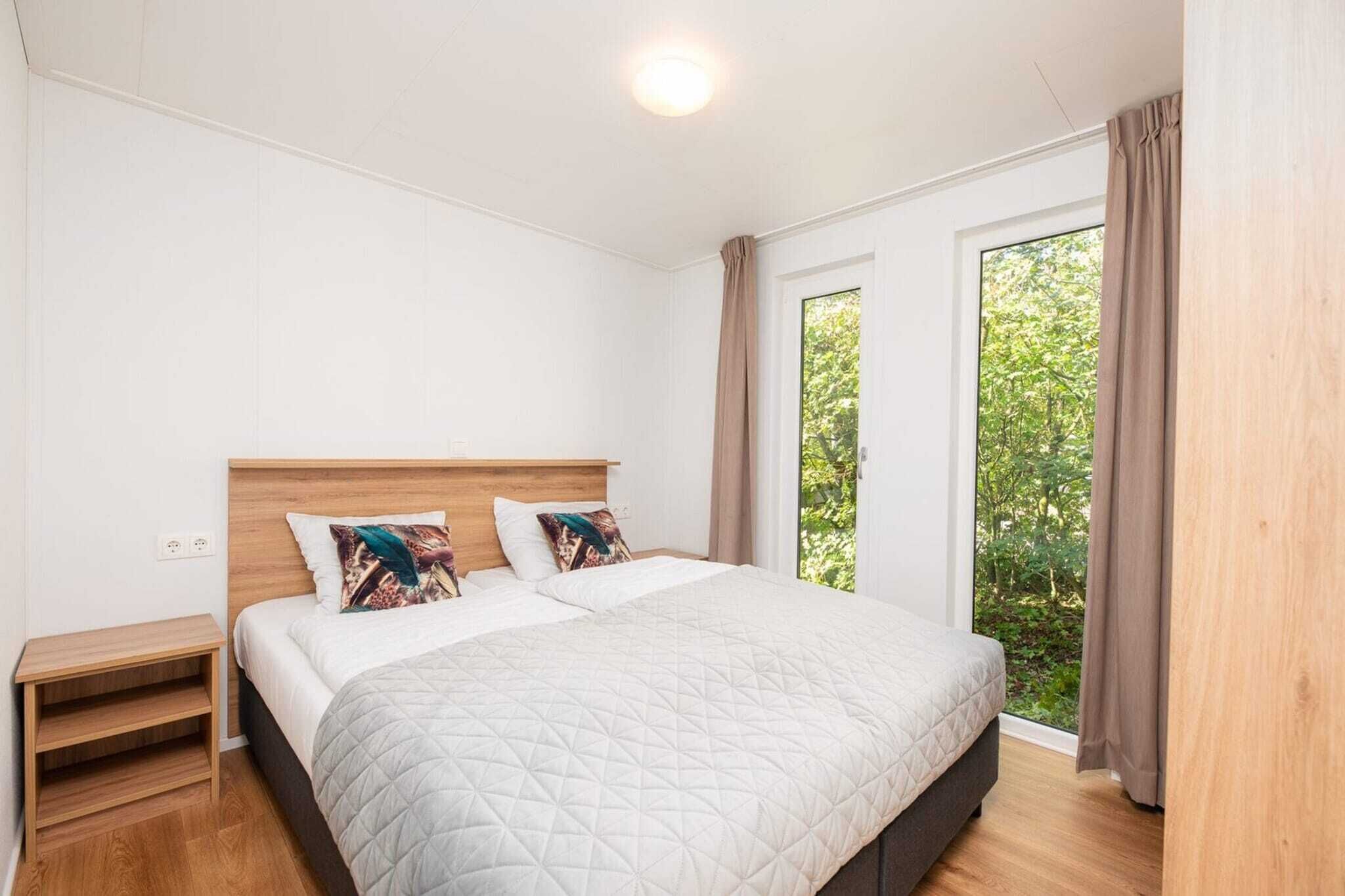 A bedroom with a bed, two pillows, a nightstand, a window with curtains, and a view of greenery.