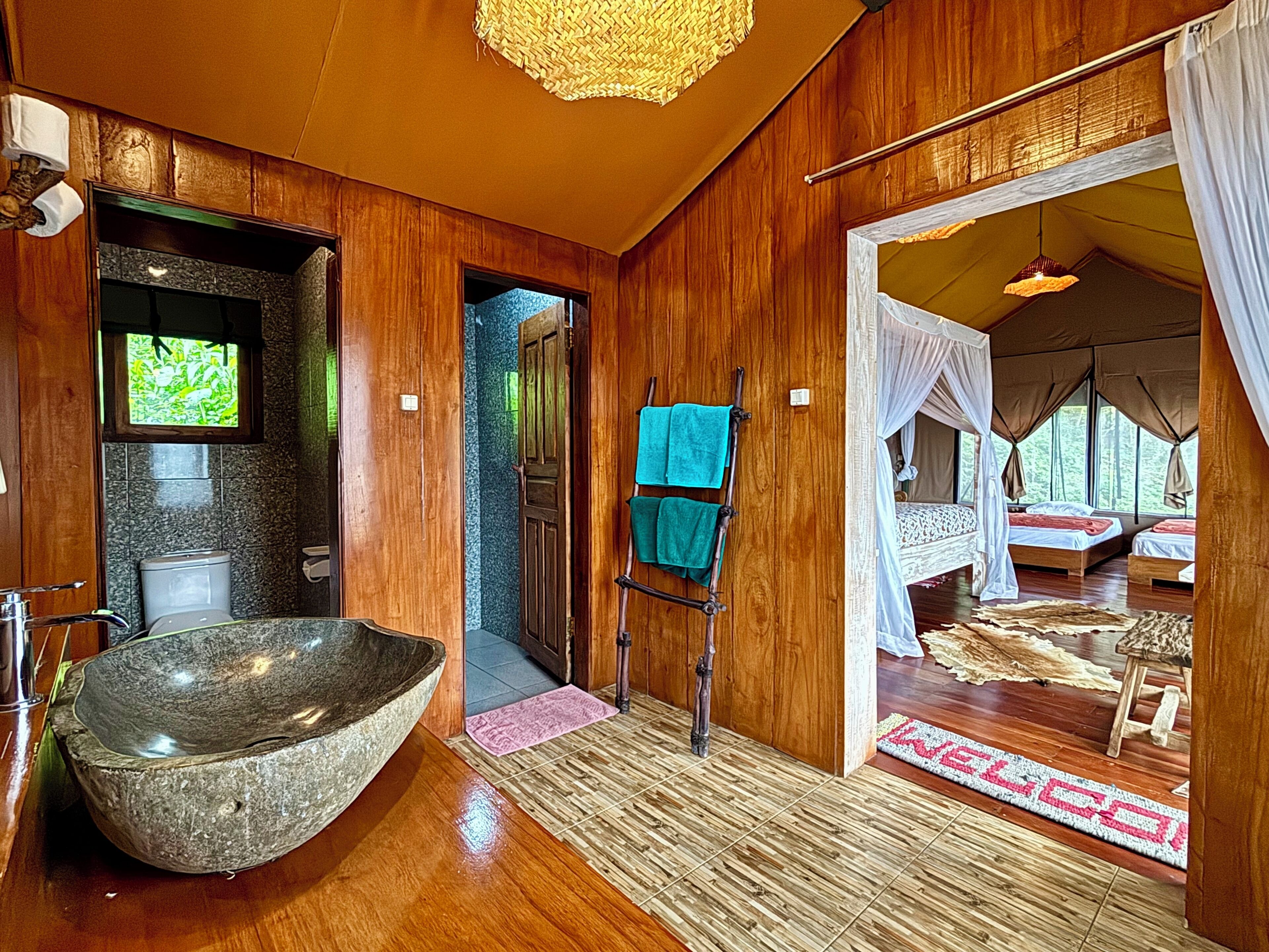 Un intérieur en bois avec un grand lavabo en pierre, une porte en bois donnant sur une chambre à coucher dotée d’un lit à baldaquin, et une vue sur une forêt par la fenêtre.