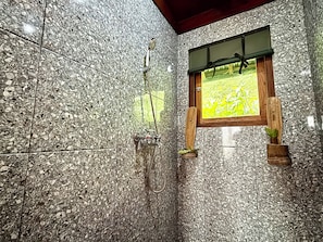 A bathroom with a granite tile wall, a shower with a hose, and a window with a view of a field.