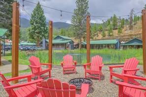 A lakeside seating area with red Adirondack chairs, a fire pit, and a view of cabins and a mountain range. - Mtn-View Red River Home & Apt Near Ski Lifts (Red River)