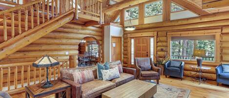 A spacious log cabin interior with a wooden staircase, leather furniture, and large windows.