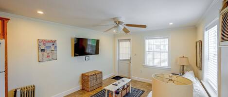 A living room with a ceiling fan, a flat-screen TV mounted on the wall, a wicker basket, a small table with magazines, and a lamp.