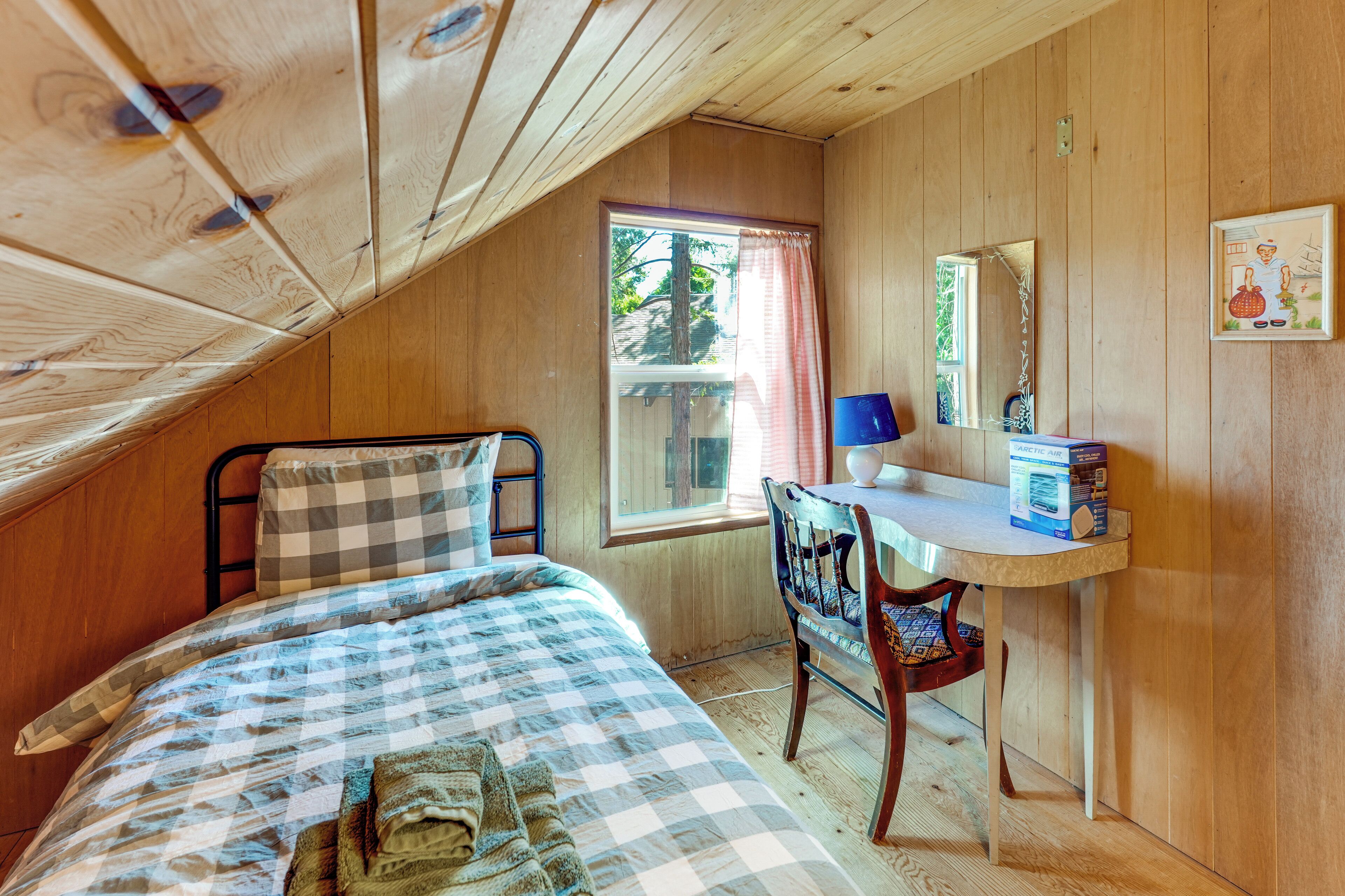 A room with a sloped ceiling, a bed with a checkered bedspread, a bedside table with a lamp, a chair, and a window with curtains.