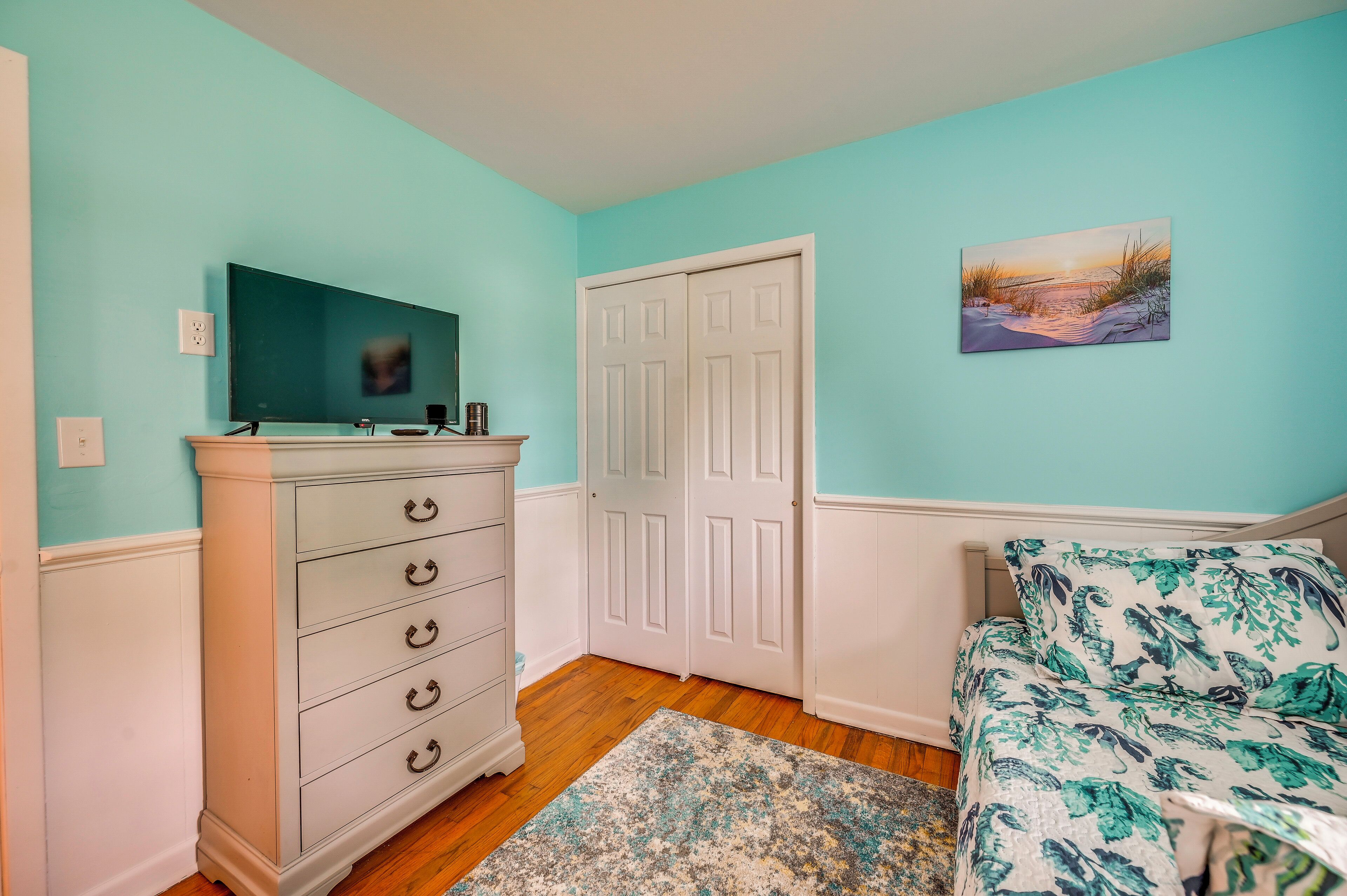Une chambre à coucher avec un lit, une commode, une porte, un tableau et un téléviseur accroché au mur.