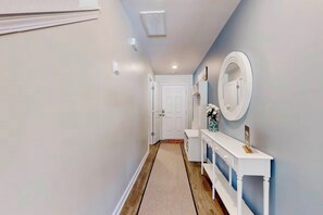 A hallway with a white door at the end, a white console table with a mirror and a vase of flowers, and a wooden floor. - Family Home w/ Game Room, Near Lake Hartwell! (Anderson)