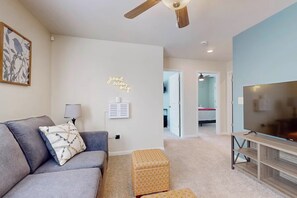 A modern living room with a grey sofa, a patterned ottoman, a ceiling fan, and a TV on a wooden stand. - Family Home w/ Game Room, Near Lake Hartwell! (Anderson)