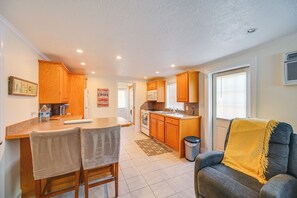A compact kitchen with wooden cabinets, a dining area with two chairs, a sofa with a yellow throw, and a window with blinds. - 2 Mi to Beach! Naples Park Bungalow (Naples)