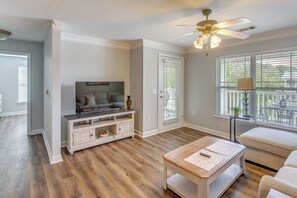A living room with a television, a ceiling fan, a window with blinds, and a wooden floor. - Golf Days, Pool Vibes! Condo 8 Mi to Sunset Beach (Calabash)