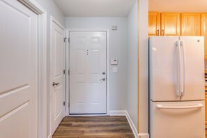 A narrow hallway with a white door at the end, a white refrigerator on the right, and wooden cabinets on the left. - Golf Days, Pool Vibes! Condo 8 Mi to Sunset Beach (Calabash)
