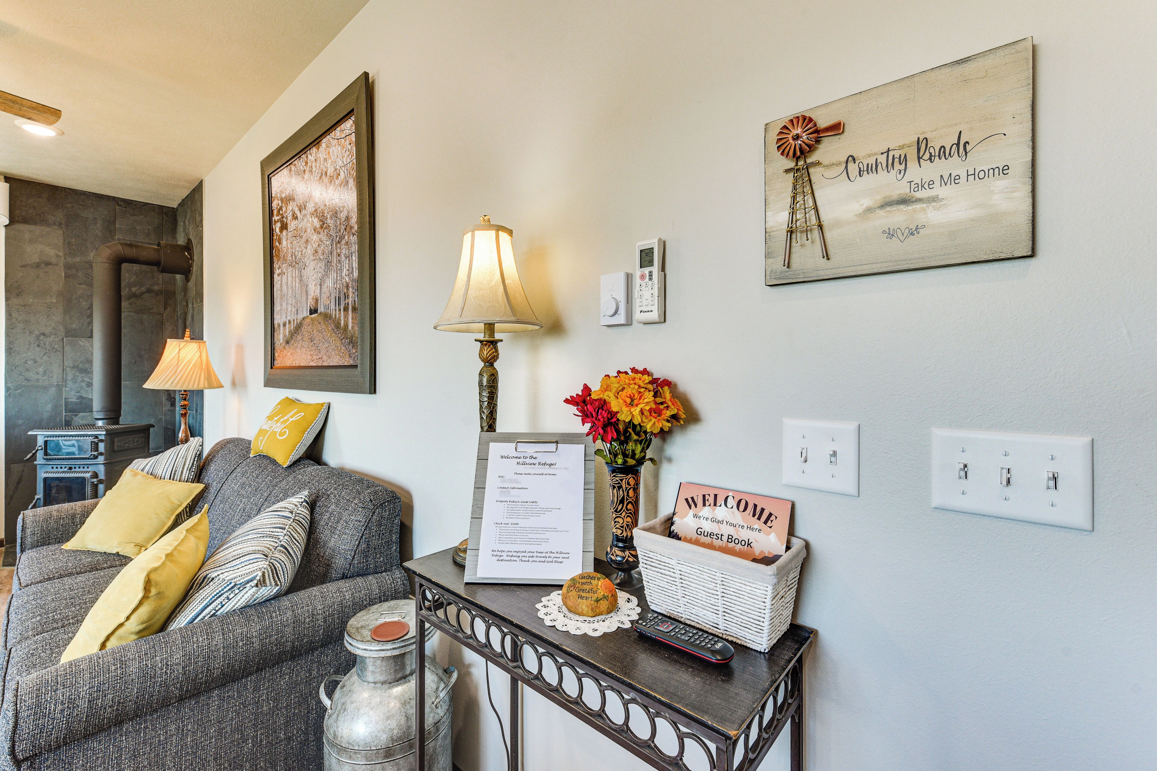 A cozy living room with a fireplace, a sofa, a table with a lamp, a wall clock, and a welcome sign.