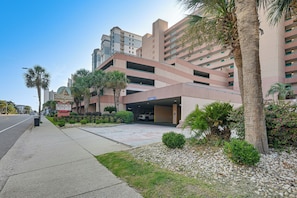 A multi-story parking garage with a clear entrance, surrounded by palm trees and a landscaped area with gravel and small shrubs. - 14th-Floor Ocean View + Pools! Myrtle Beach Haven (Myrtle Beach)