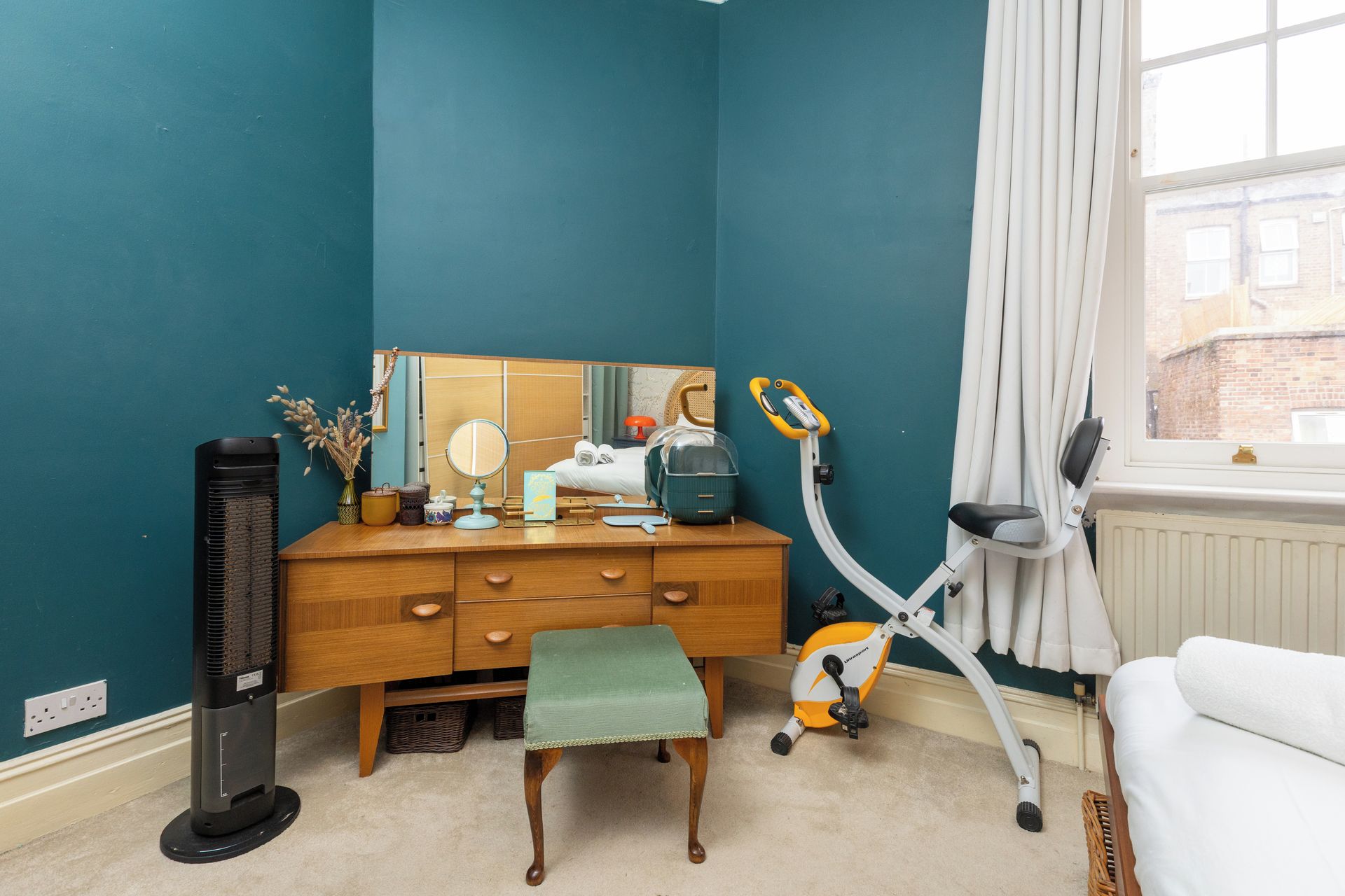 A room with teal walls, a wooden dresser, a bench, a standing heater, a mirror, a suitcase, and a window with curtains.