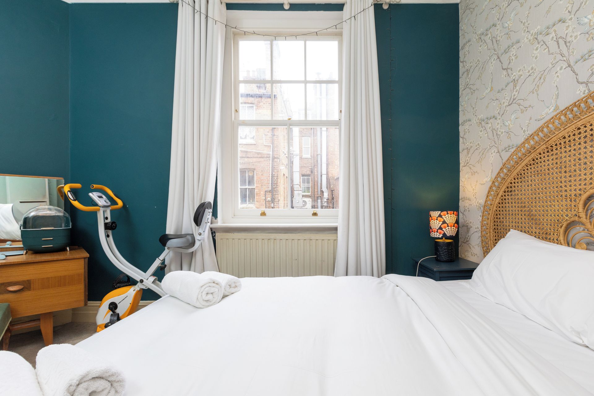 A hotel room with a bed, a window with curtains, a desk with a lamp, and exercise equipment.