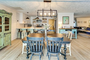 Un coin repas avec une table et des chaises en bois, une cuisine à l’arrière-plan et un salon avec un canapé et un téléviseur.