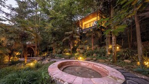 A circular stone hot tub surrounded by rocks and plants, with a wooden bridge and a building with a thatched roof in the background.